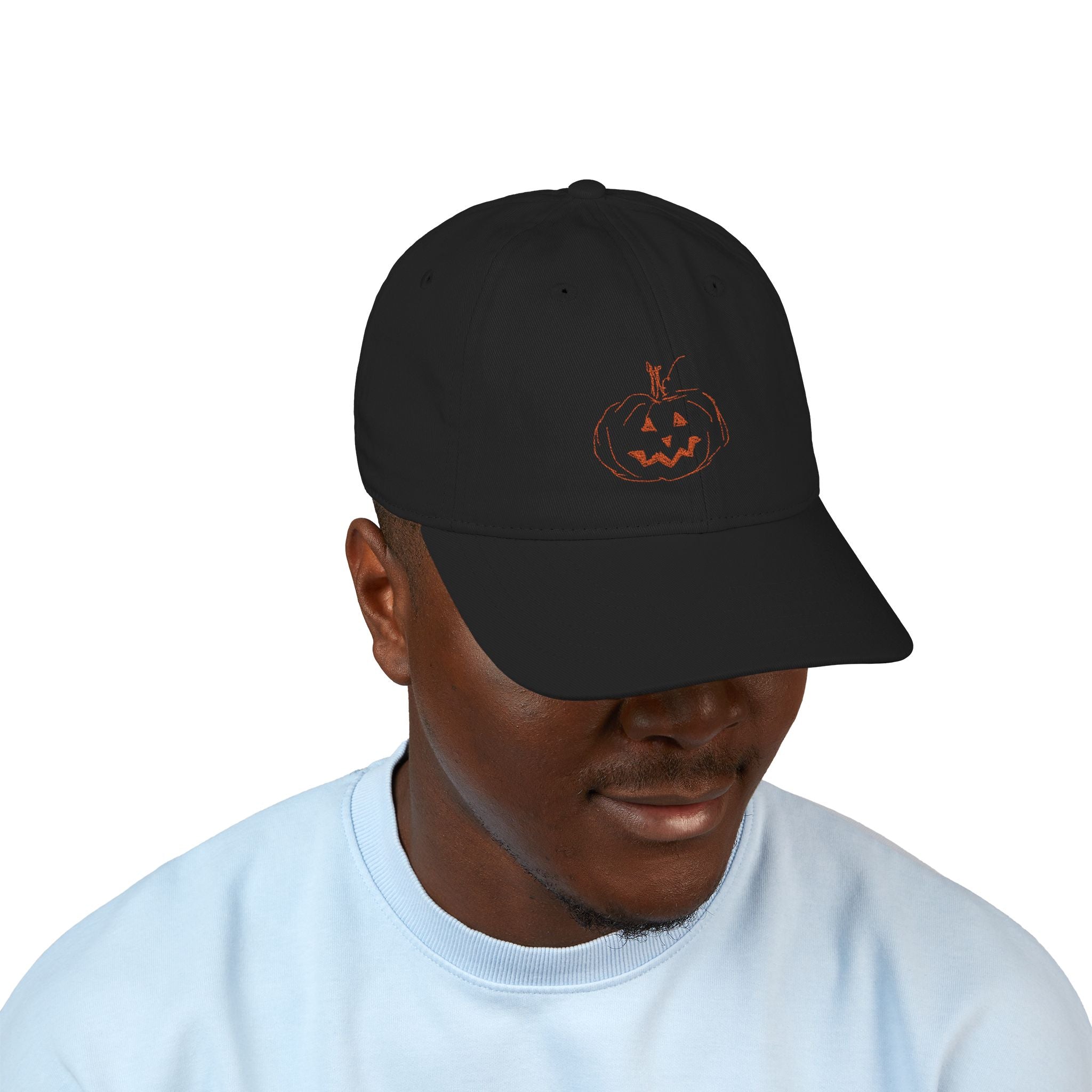 Male wearing a black baseball cap with embroidered jack-o-lantern pumpkin outline design on the front in a rusty orange color.