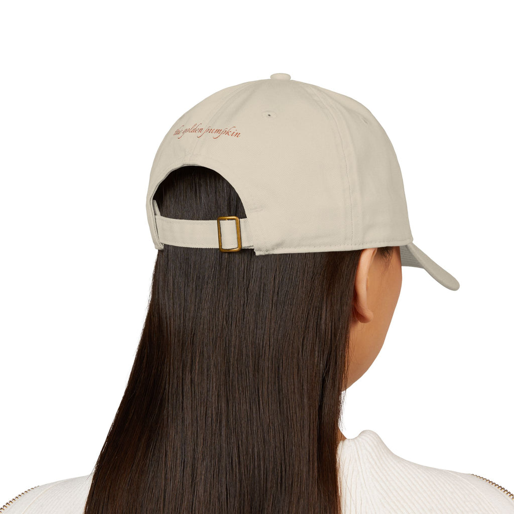 Female wearing a beige baseball cap with text 'the golden pumpkin' on the back, in rusty orange color. The golden pumpkin is the store name.